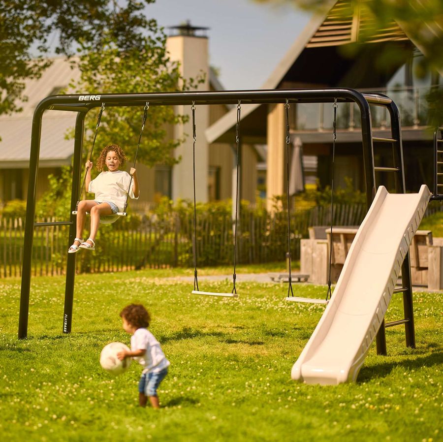 Child on a swing seat on a BERG PlayBase frame fitted with a trapeze and a slide
